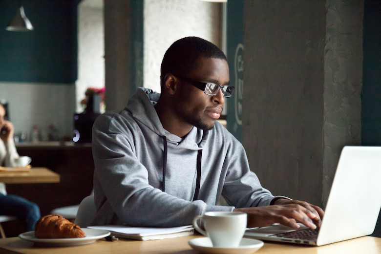 Serious millennial african-american man using laptop writing emails, distantly working or studying on computer in public place